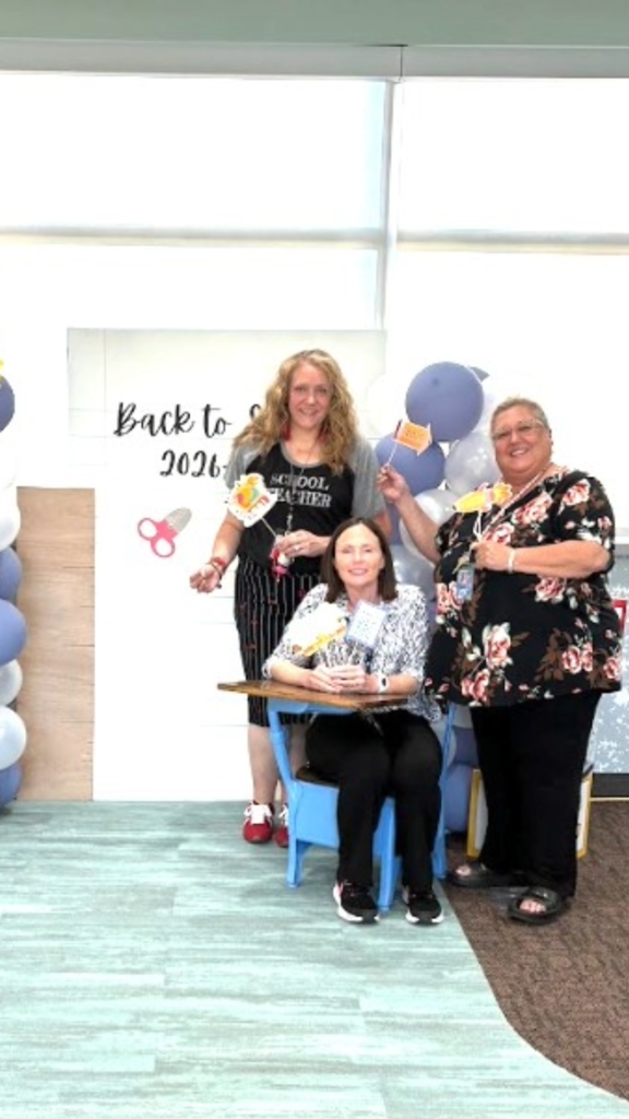 Three women posing with photo booth props in front of a backdrop reading "Back to School 2026-2027". A woman on the left wears a shirt reading "School Teacher" and holds a scissor prop, a woman in the center sits at a blue desk holding classroom props, and a woman on the right stands holding a pencil prop.