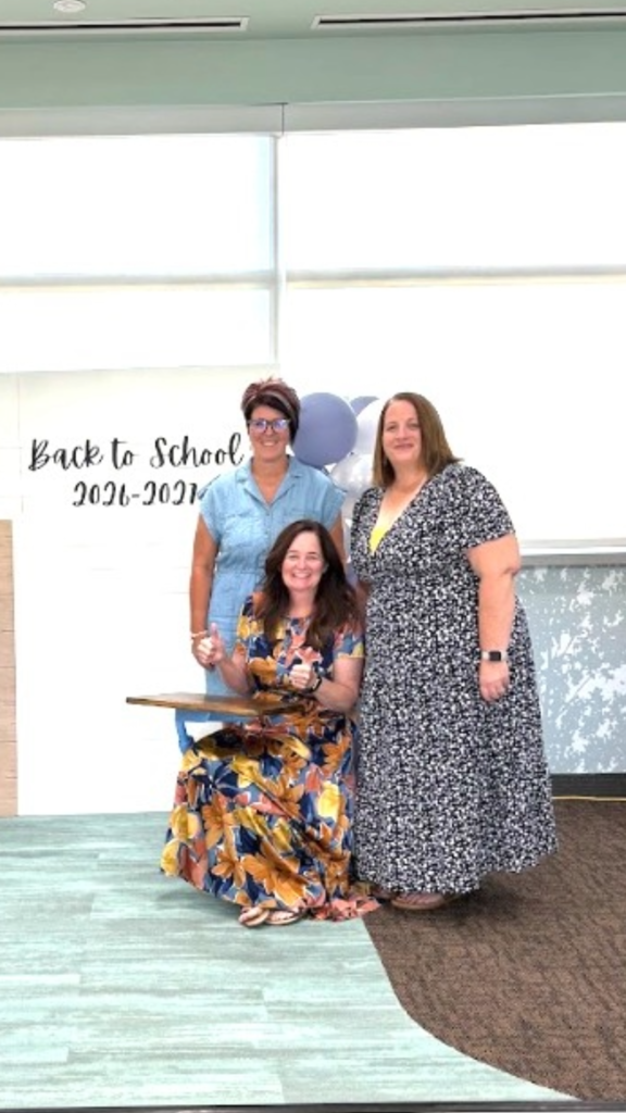 Three women posing in front of a backdrop displaying "Back to School 2026-2027" and blue and white balloons. A woman in a denim dress stands on the left, a woman in a colorful floral dress sits at a wooden school desk in the center giving two thumbs-up gestures, and a woman in a black-and-white patterned dress stands on the right.