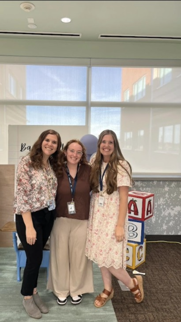 Three women standing side-by-side and smiling in front of a "Back to School" backdrop framed by large ABC toy blocks on the right. The woman on the left wears a floral patterned blouse and black trousers, the woman in the center wears a brown top and wide-leg tan pants, and the woman on the right wears a cream floral lace dress with brown strap sandals.