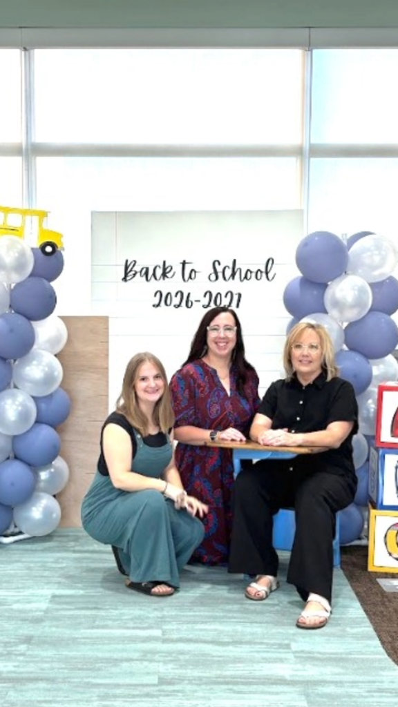 Three women posing in front of a white backdrop displaying the text "Back to School 2026-2027" flanked by blue and white balloon arches with a yellow school bus cut-out on top. One woman in a teal jumpsuit crouches on the left, a woman in a red paisley dress stands in the center, and a woman in a black outfit sits at a small blue school desk on the right next to stacked ABC blocks.