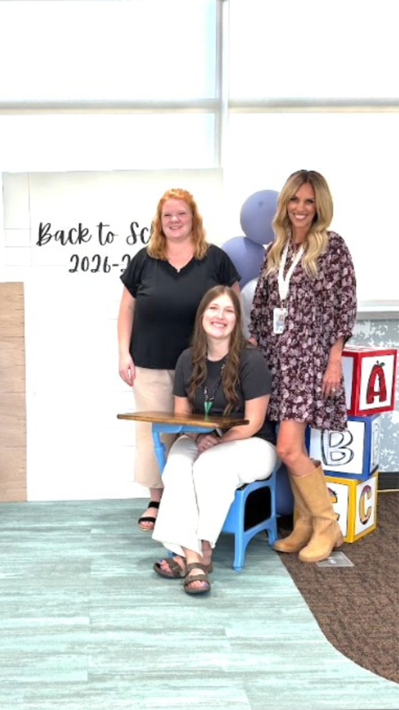 Three women smiling near a blue school desk against a white backdrop reading "Back to School 2026-2027" next to large ABC building blocks. A woman in a black V-neck shirt and tan pants stands on the left, a woman in a gray shirt and white pants sits at the desk, and a woman in a dark floral dress and tan boots stands on the right.