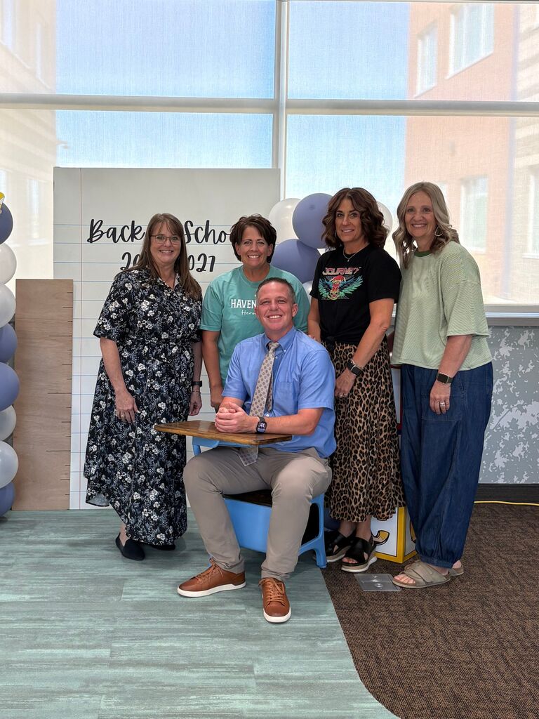 Five staff members posing in front of a backdrop reading "Back to School 2026-2027". A man in a blue button-down shirt, tie, and tan pants sits at a small blue school desk in the center, surrounded by four women in casual and patterned outfits standing in a row behind him.