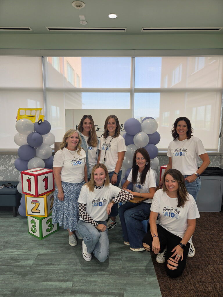 Seven female school staff members wearing matching white t-shirts that read "Haven Bay AIDE" pose together in front of a "Back to School" balloon display and oversized alphabet blocks.
