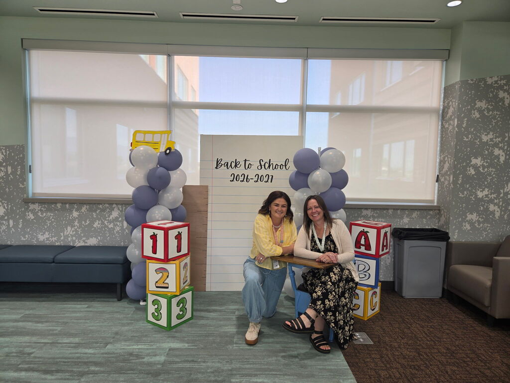 Two female staff members pose at a "Back to School 2026-2027" photo backdrop lined with purple and white balloon towers, large toy blocks showing numbers and letters, and a giant sheet of notebook paper displaying the text "Back to School 2026-2027".