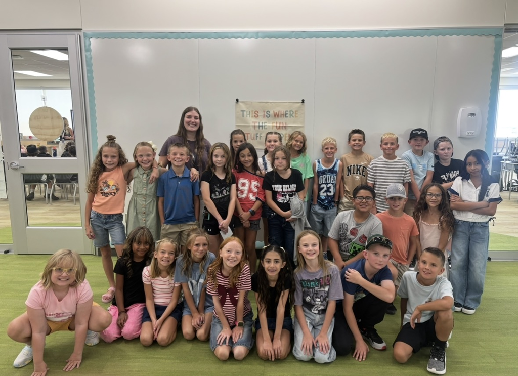 A group of elementary school students poses inside a classroom with their teacher under a small cloth sign on the wall that reads "THIS IS WHERE THE FUN STUFF HAPPENS.
