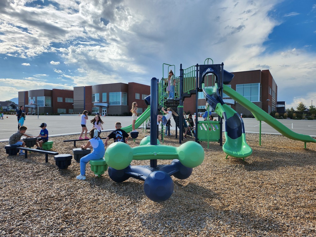 Children play on a green and blue playground structure situated on a wood-chip surface outside a modern brick school building under a cloudy sky.