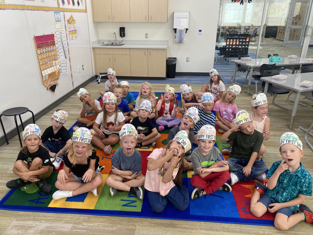 A group of young kindergarten students sit on a colorful alphabet rug wearing paper crown hats that read "I'm in K - HAPPY FIRST DAY" while making fun faces for the camera.