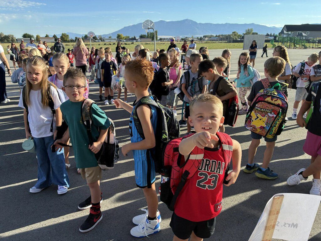 Young elementary students line up outdoors on the school blacktop wearing backpacks, with a boy in front wearing a red "JORDAN 23" shirt pointing at the camera, and a sign in the lower right partially reading "MRS..."