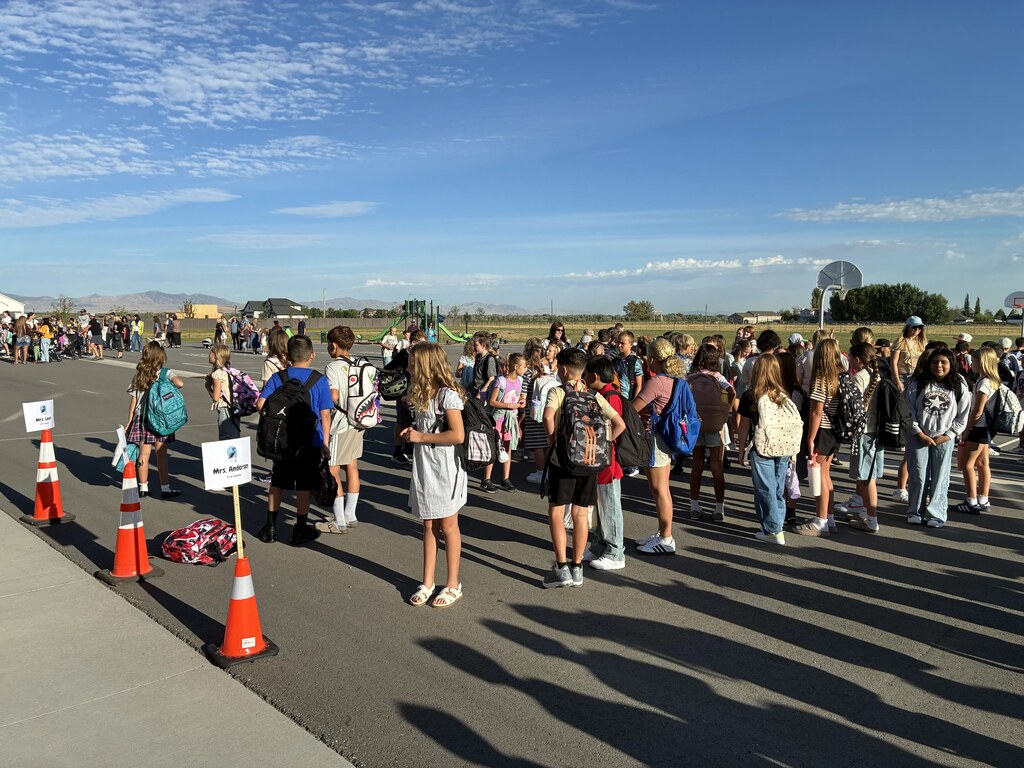 Elementary students gather in organized lines outside on the school asphalt on a sunny morning next to orange traffic cones holding signs labeled "Mrs. Lee" and "Mrs. Anderson".