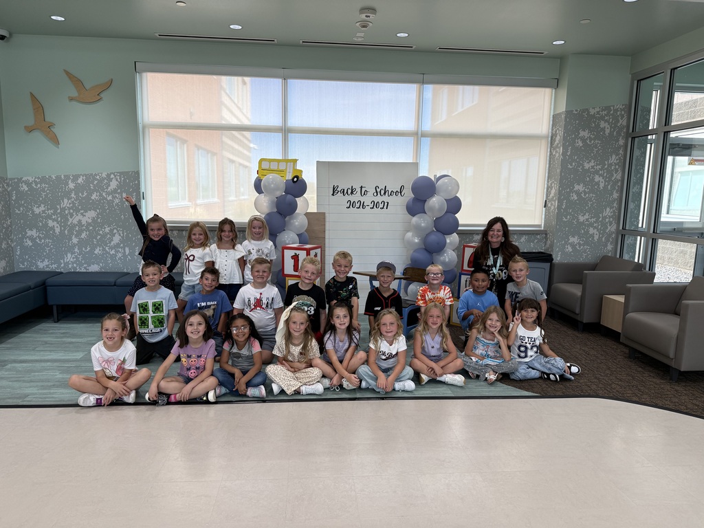 A large class of young students and their teacher pose indoors on a colorful rug in front of a "Back to School 2026-2027" photo backdrop featuring balloon columns and giant numbered blocks.