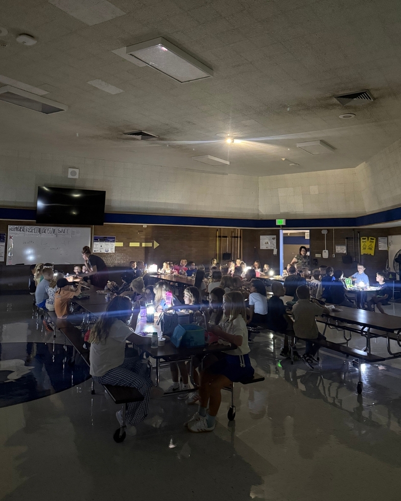 A dimly lit school cafeteria filled with elementary-aged students seated at long tables during an evening event. Students hold small lights or glow sticks, creating a bright, glowing atmosphere, while several adults supervise throughout the room. A whiteboard and wall-mounted television are visible in the background.