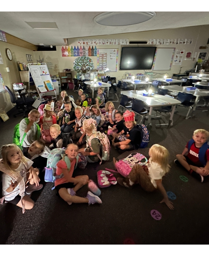 A group of elementary school students sit together on the classroom floor, smiling for a photo. They wear backpacks and colorful clothing, and one student wears a red chef hat. Desks, chairs, classroom displays, a whiteboard, and a large screen are visible in the background.