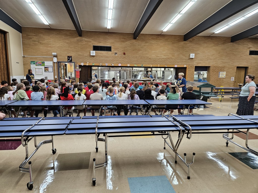 A group of elementary students sitting on the cafeteria floor listening attentively to staff members explaining cafeteria rules and expectations.