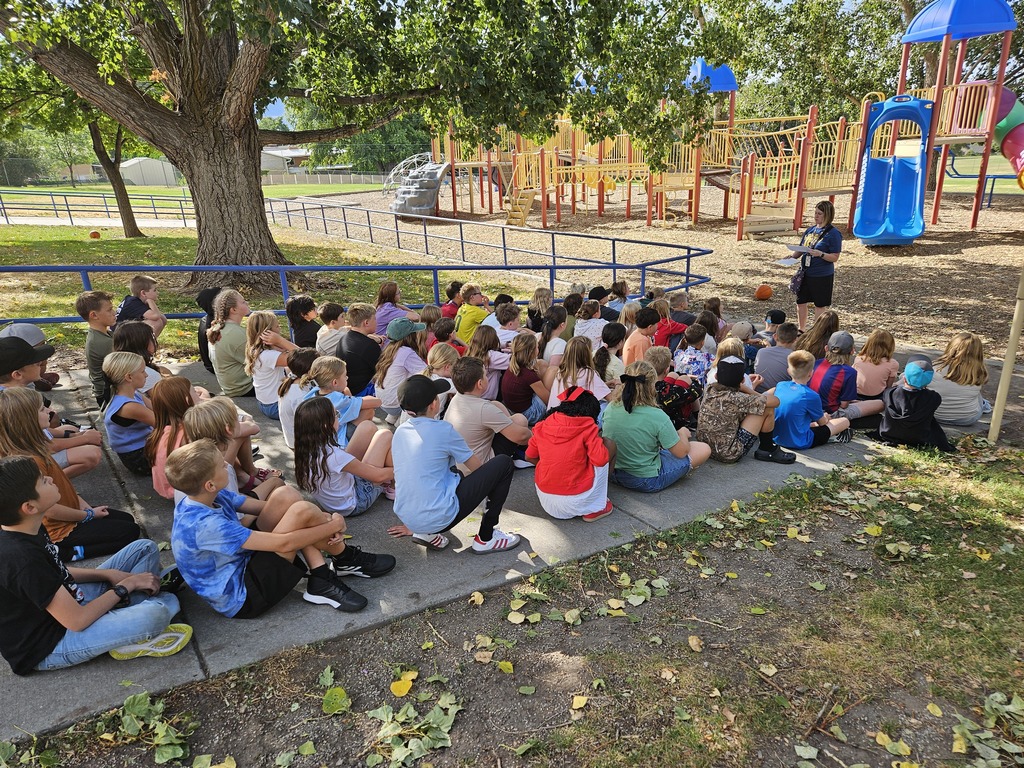 Elementary students sitting together on the concrete near a shaded playground area listening to a teacher outline playground procedures.