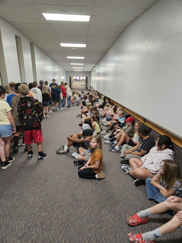 Elementary students sitting quietly along the wall in a carpeted school hallway while a staff member addresses them.