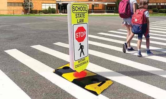 An in-street pedestrian crosswalk safety sign mounted on a black and yellow weighted base placed on white zebra crosswalk stripes. Two young students wearing backpacks walk in the background. Sign text reads "SCHOOL - STATE LAW - STOP - WITHIN CROSSWALK" with a red octagon stop symbol and a black pedestrian icon.