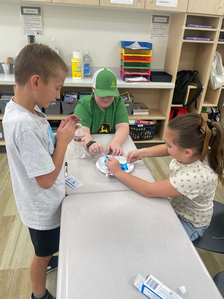 Three students collaborating over a paper plate with toothpaste at joined classroom desks, with one student wearing a green John Deere hat and t-shirt featuring the John Deere logo. Background text on classroom wall signs and book covers includes "Turn In Procedures", "Respect each other", "GHOSTS", and "EVERYDAY".