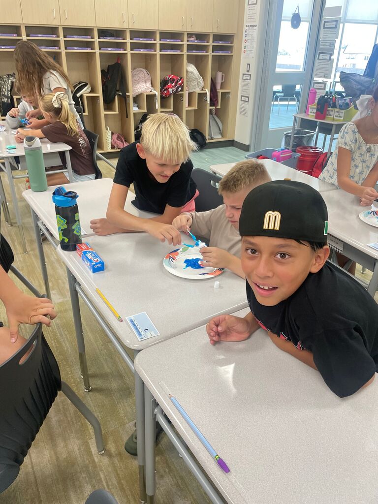 Three students sitting at classroom desks working together on a toothpaste activity using a paper plate, with Crest Cavity Protection toothpaste on the desk. One student in a black hat with a yellow "M" logo smiles at the camera in the foreground. Labels visible on posters in the background include "IN AN EMERGENCY" and "SOAR".