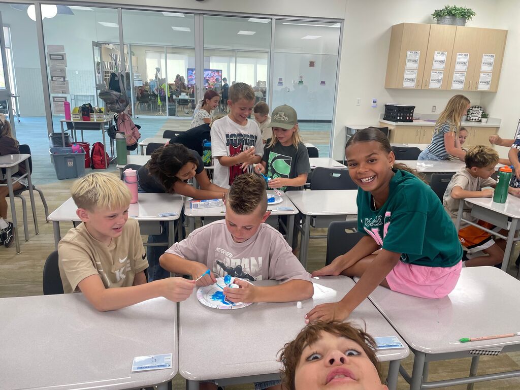 A classroom full of students engaging in a hands-on desk activity with blue toothpaste, paper plates, and cotton swabs. A student leaning in from the bottom right makes a humorous face toward the camera. Visible clothing text includes "JORDAN" on a student's white t-shirt and "Nike" on a tan shirt.