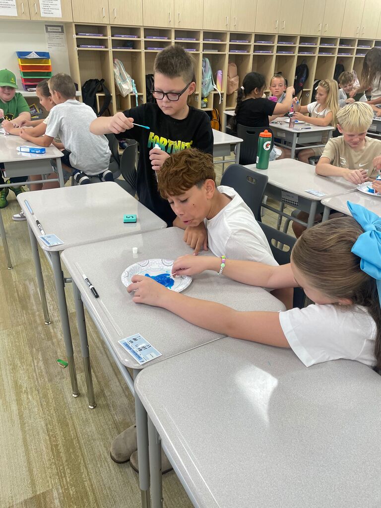 tudents seated at classroom desks working on a group activity using cotton swabs, blue paste, and paper plates. A student standing in the middle wears a black t-shirt with "FORTNITE" printed in yellow text. Desk labels show numbers "5" and a name card reading "Lakes Elementary S.O.A.R.".
