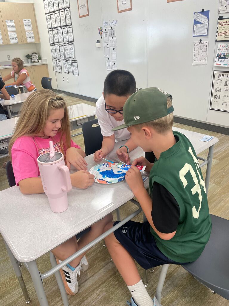 Three students working together around a paper plate containing blue toothpaste at a classroom desk. A pink Hydro Flask water bottle sits on the left, and a student on the right wears a green jersey with the number "31". Background signs on the wall include "Science", "fast finisher", "A Teacher's Top Secret", "CONFIDENTIAL", "A Letter From Your Teacher", and "HAPPY BIRTHDAY".