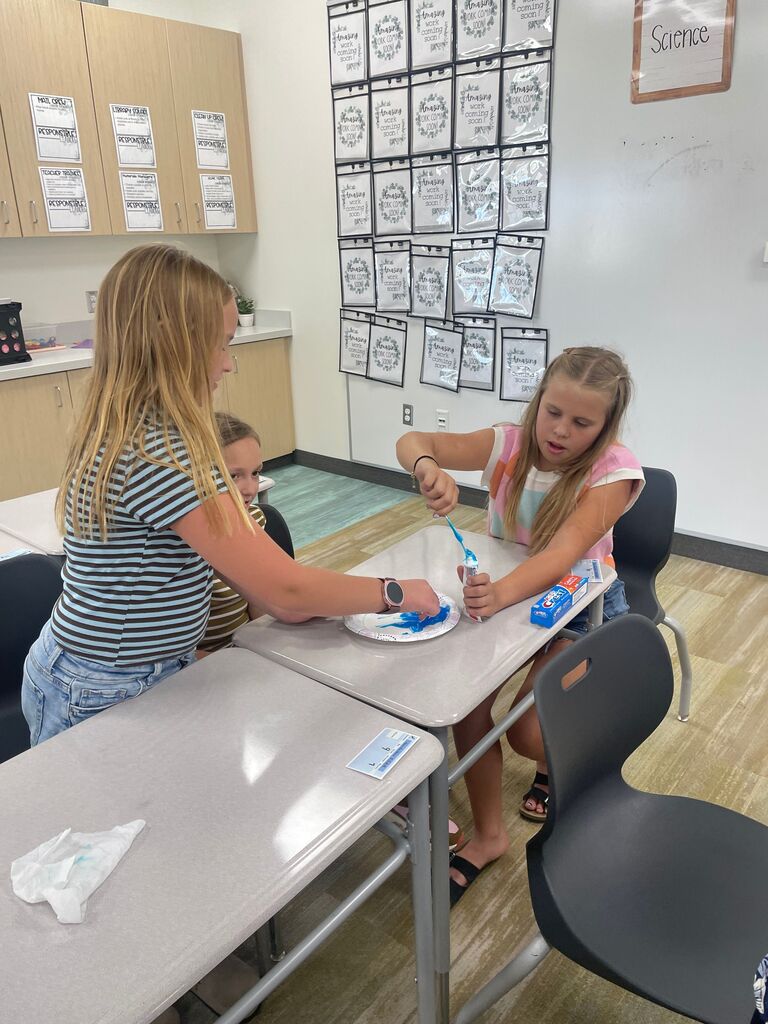 Elementary school students standing and sitting around classroom desks during a hands-on activity with blue toothpaste and a paper plate. Visible background text includes "Science" on a classroom wall sign, "MAT CREW", "LIBRARIAN ASSISTANT", "CLEAN UP CREW", "TEACHER ASSISTANT", "MATERIALS MANAGER", "SOAR TEAM", "RESPONSIBLE", and multiple wall pockets labeled "Amazing work coming soon!".