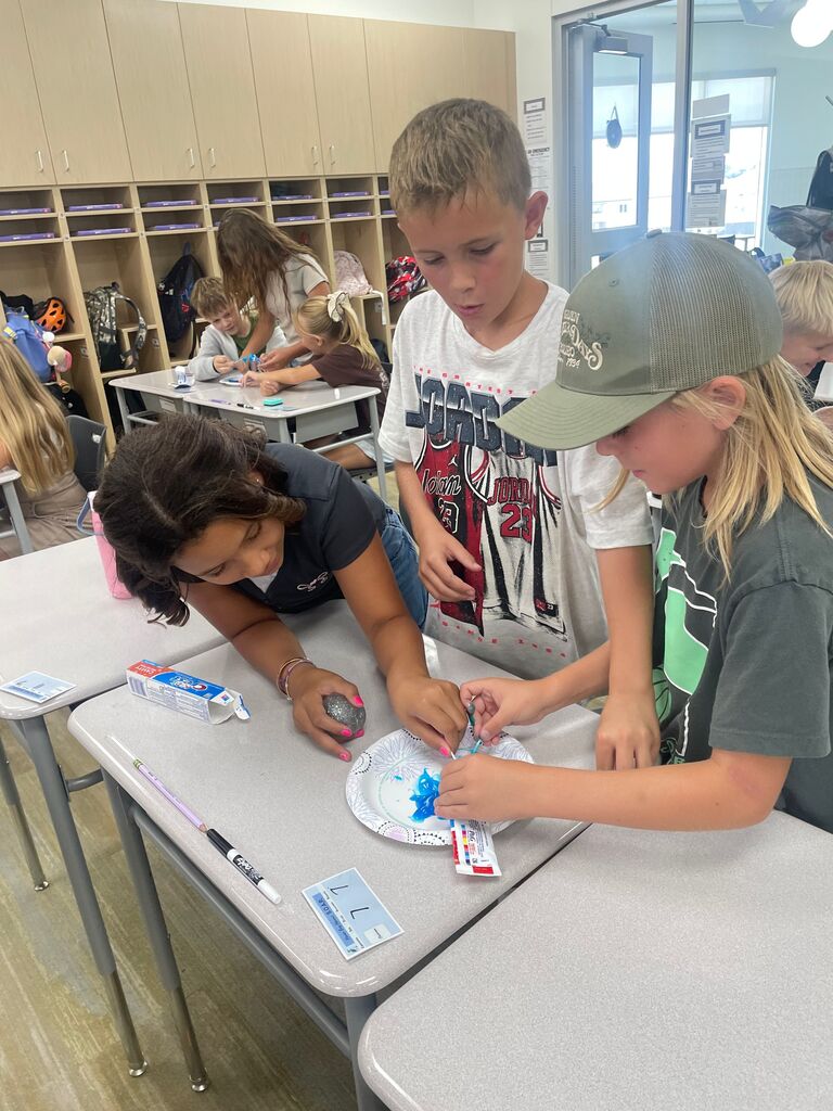 Three students gathered around a classroom desk during a toothpaste science experiment using a paper plate and a Crest Cavity Protection box. The middle student wears a white graphic shirt with the word "JORDAN" printed across the front, and the student on the right wears a green baseball hat featuring the text "EST. 1994". A small desk tag displays "7" with "Lakes Elementary S.O.A.R.".