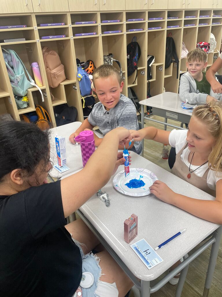 Three students sitting at a classroom desk experimenting with squeezing blue toothpaste from a tube labeled "Crest CAVITY" onto a paper plate. A blue label on the desk displays "14" alongside "Lakes Elementary S.O.A.R.".