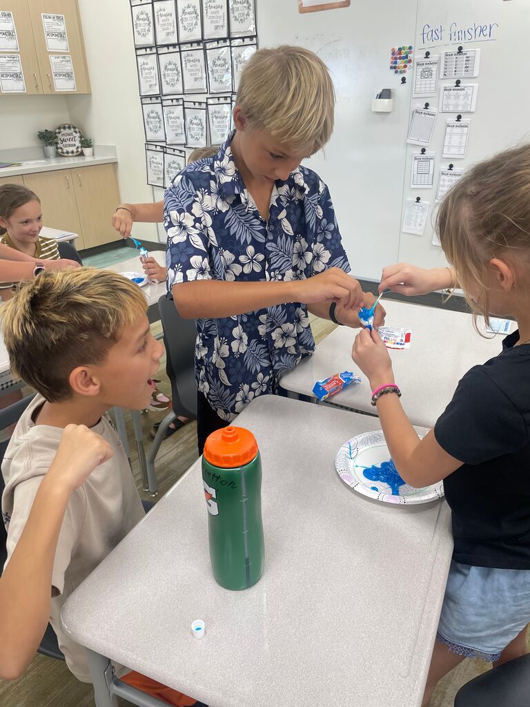 A boy wearing a blue floral shirt and a girl working together to squeeze blue toothpaste onto a paper plate while another student watches enthusiastically. Posters in the background feature text including "fast finisher", "RESPONSIBLE", and repeated prints of "Amazing work coming soon!".