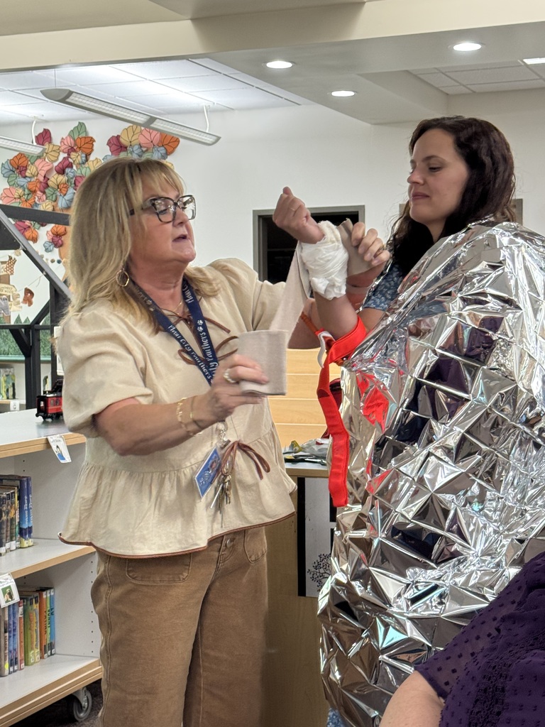 Becci helping Miss Dawna wrap bandages on her arm. Miss Dawna is wearing a reflective blanket. Together they are teaching staff about safety procedures.