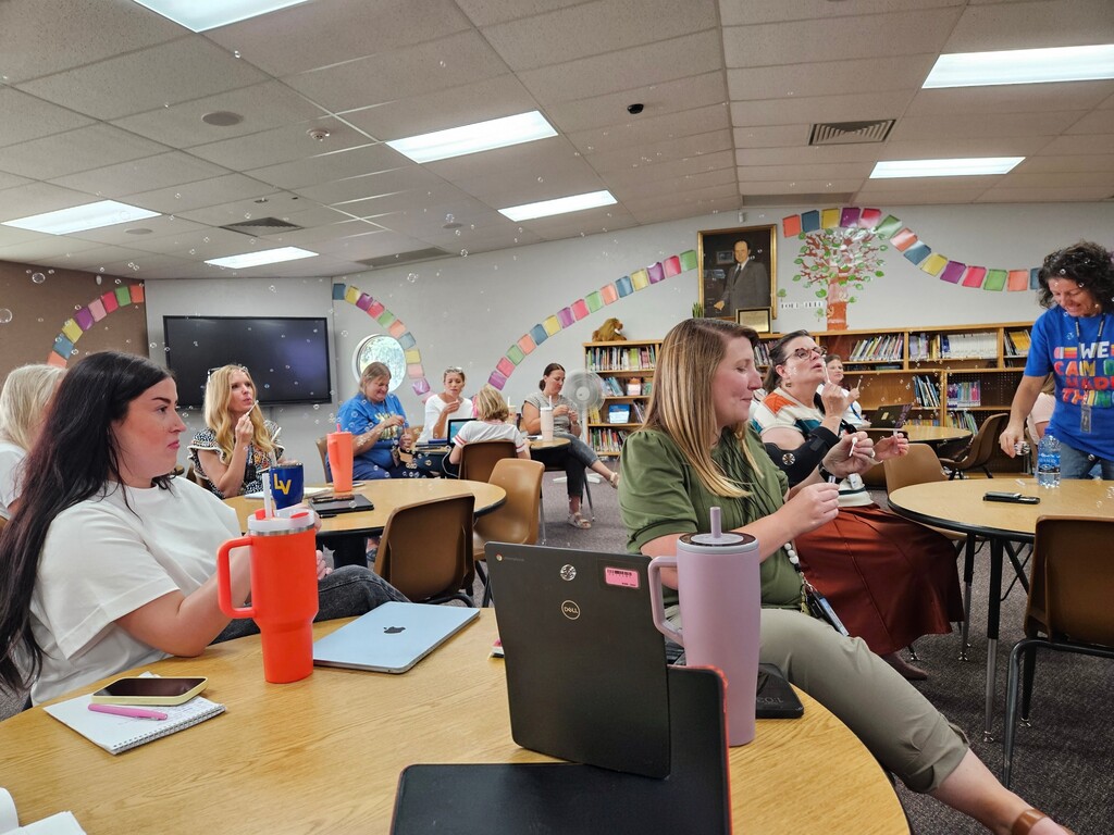 Elementary school teachers and staff sit at round tables in the school library blowing soap bubbles during an after-school meeting.