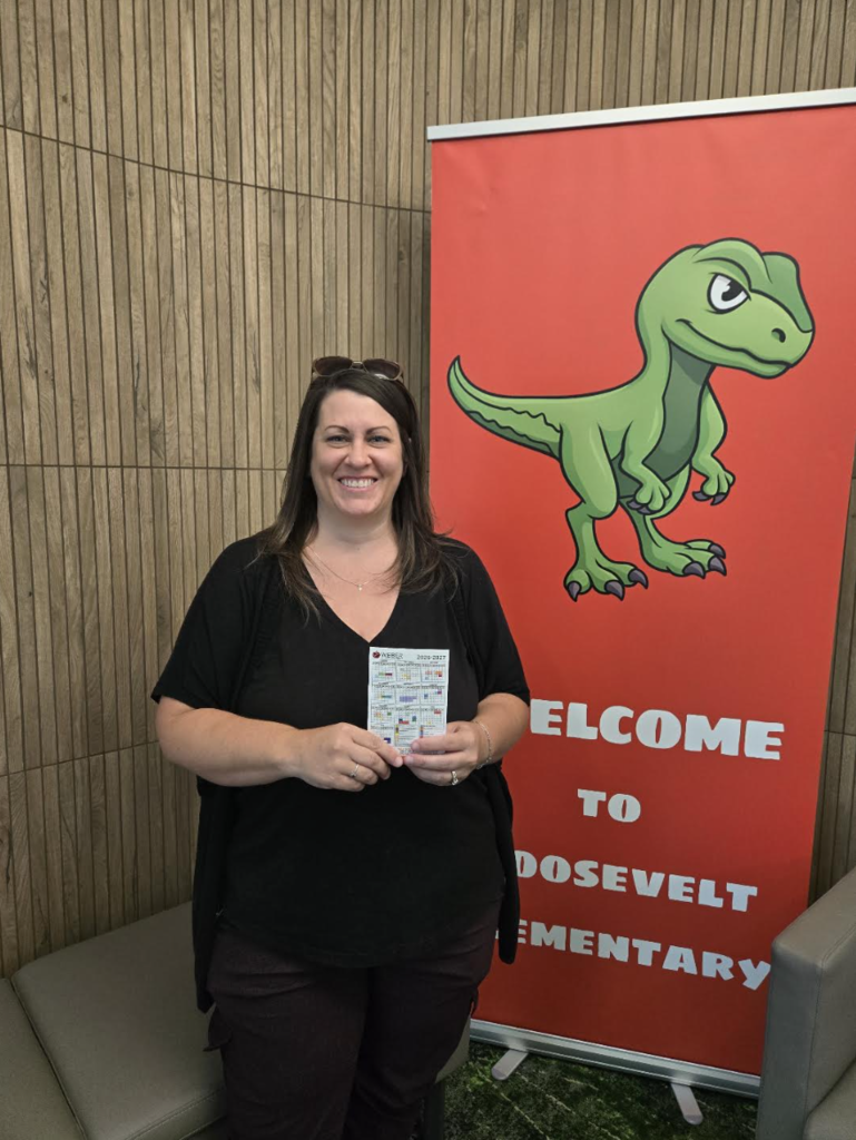 woman holding a calendar magnet posing in front of a welcome to roosevelt elementary banner