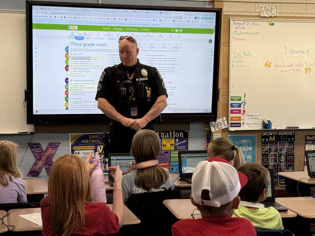 School Resource Officer Done stands at the front of a Lomond View Elementary classroom in front of an interactive display screen displaying IXL Math, introducing himself to 3rd and 4th grade students.