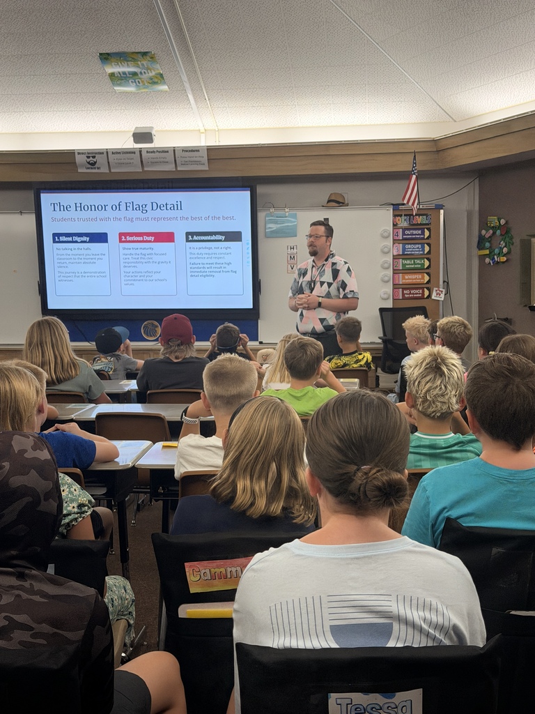 A male teacher in a patterned shirt standing at the front of a classroom presenting a digital slideshow titled "The Honor of Flag Detail" to a room full of 5th-grade students seated at their desks.