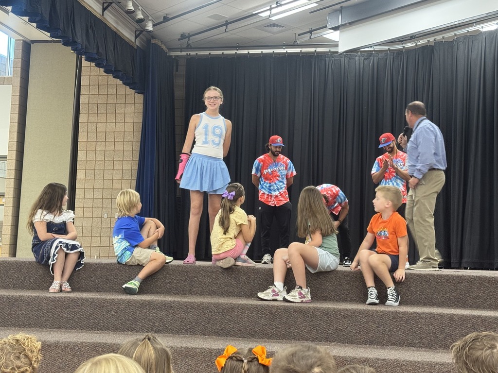 Three Ogden Raptors baseball players wearing their jerseys stand on the stage in the Plain City Elementary gymnasium, speaking into microphones to a crowd of preschool through sixth-grade students sitting on the floor.