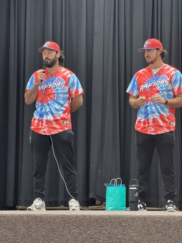 Three Ogden Raptors baseball players wearing their jerseys stand on the stage in the Plain City Elementary gymnasium, speaking into microphones to a crowd of preschool through sixth-grade students sitting on the floor.