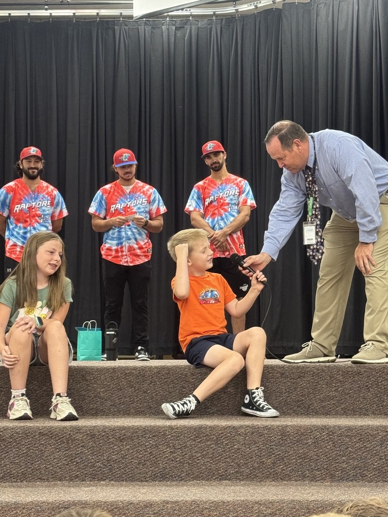 Three Ogden Raptors baseball players wearing their jerseys stand on the stage in the Plain City Elementary gymnasium, speaking into microphones to a crowd of preschool through sixth-grade students sitting on the floor.