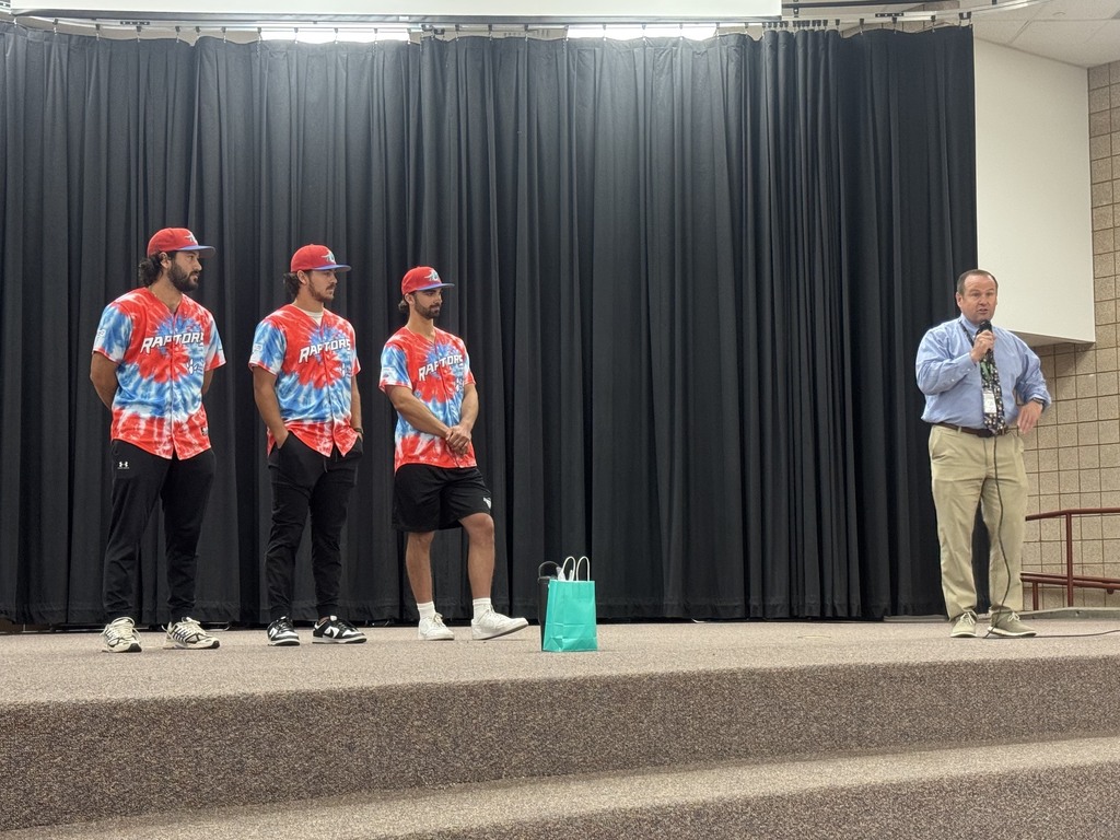 Three Ogden Raptors baseball players wearing their jerseys stand on the stage in the Plain City Elementary gymnasium, speaking into microphones to a crowd of preschool through sixth-grade students sitting on the floor.