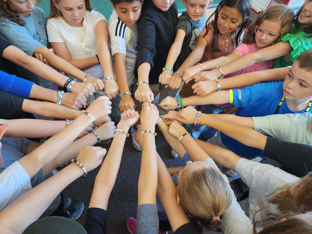 A close-up view of a group of elementary students from Mrs. Van Meeteren's class placing their hands together in the center of a circle. Each student is wearing a handmade bead bracelet spelling out the word "BELIEVE" in blue, black, and white beads.