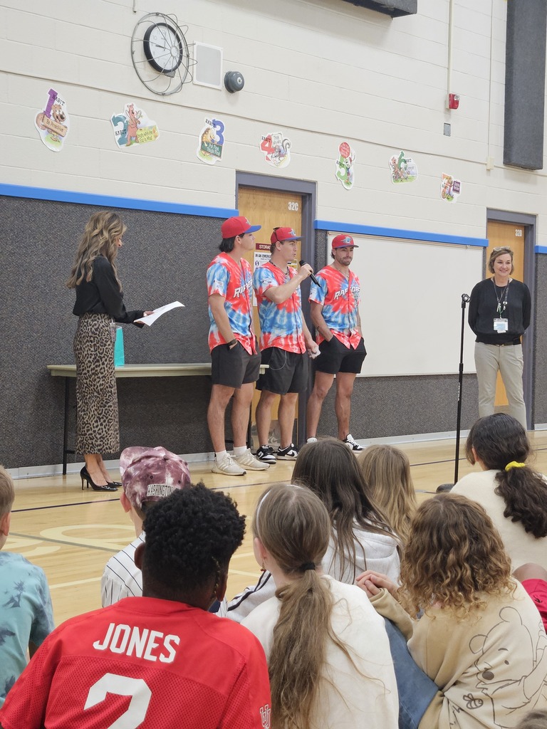 Three Ogden Raptors players in matching tie-dye jerseys standing at the front of a gym holding a microphone while students sit on the floor watching.