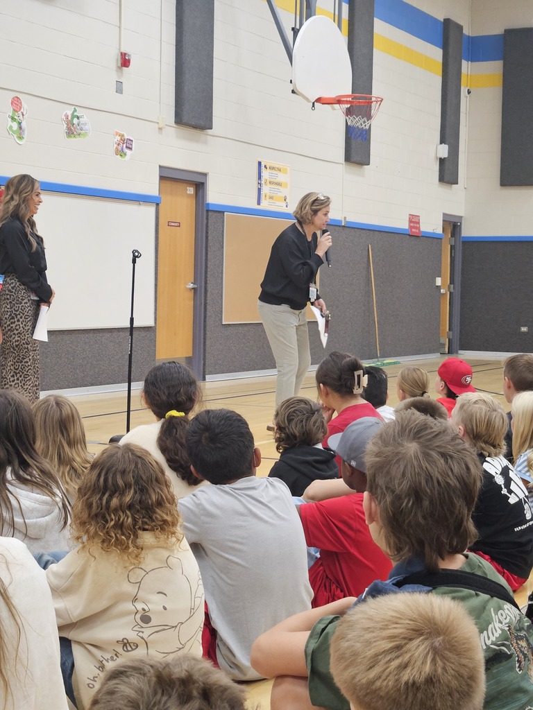 Superintendent Gina Butters, wearing a dark top and light pants, leaning down to hand a microphone to a student sitting on the gym floor during an assembly.