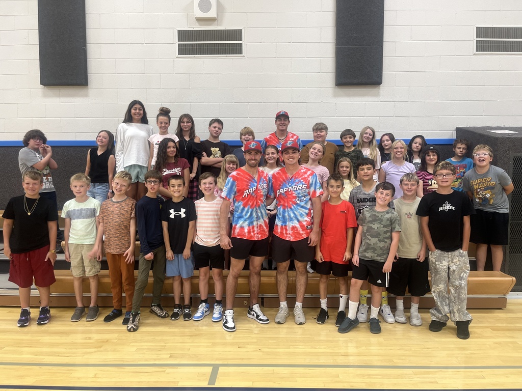 A large group of elementary students lined up in front of gym bleachers posing with two Ogden Raptors players wearing tie-dye jerseys and red caps.