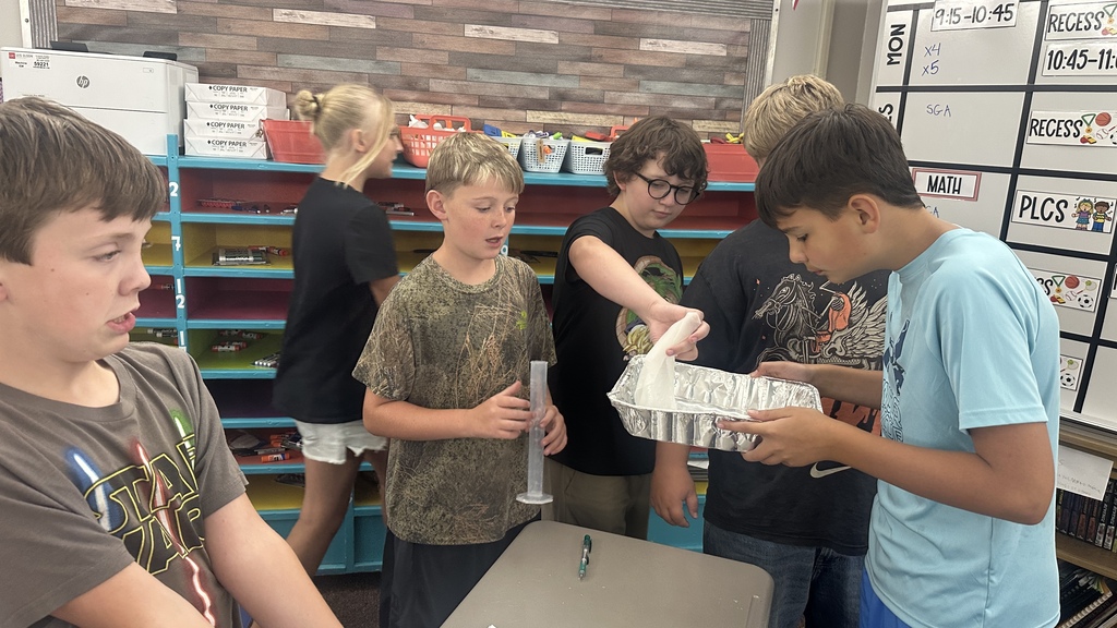 Students gathered around a desk using a clear graduated cylinder and an aluminum pan during a hands-on classroom lab. 