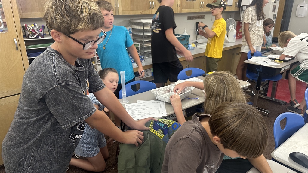 Students gathered around a desk using a clear graduated cylinder and an aluminum pan during a hands-on classroom lab. 