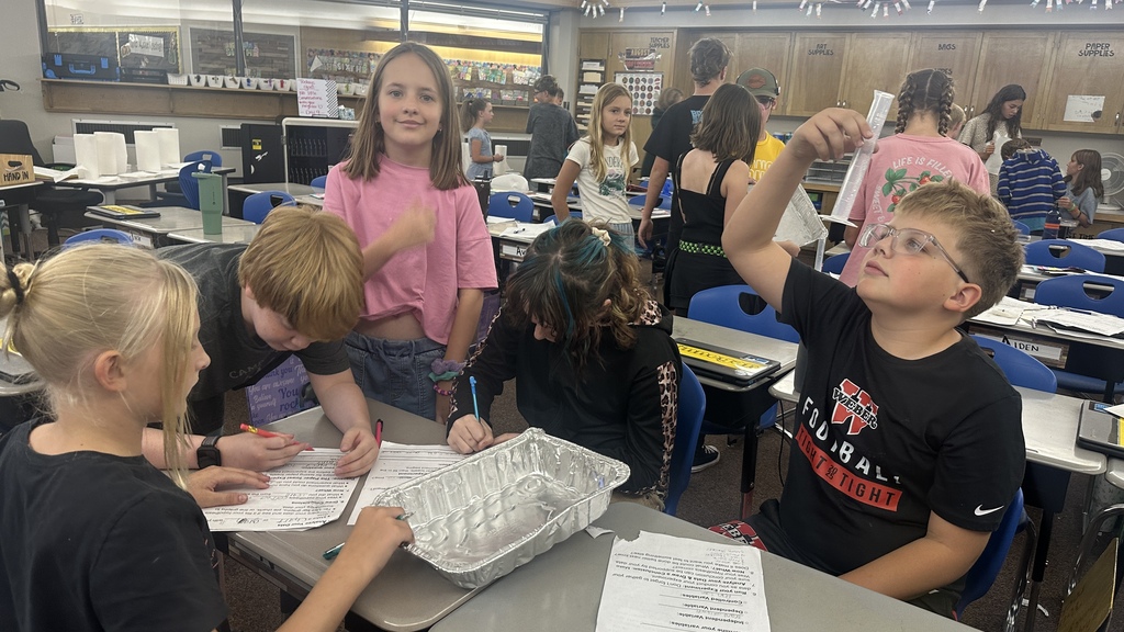 Students gathered around a desk using a clear graduated cylinder and an aluminum pan during a hands-on classroom lab. 