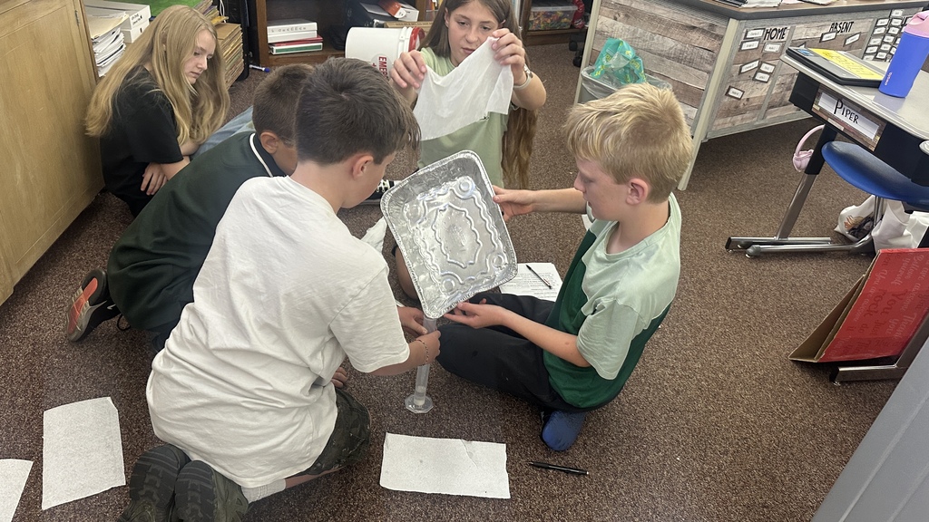 Students gathered around a desk using a clear graduated cylinder and an aluminum pan during a hands-on classroom lab. 