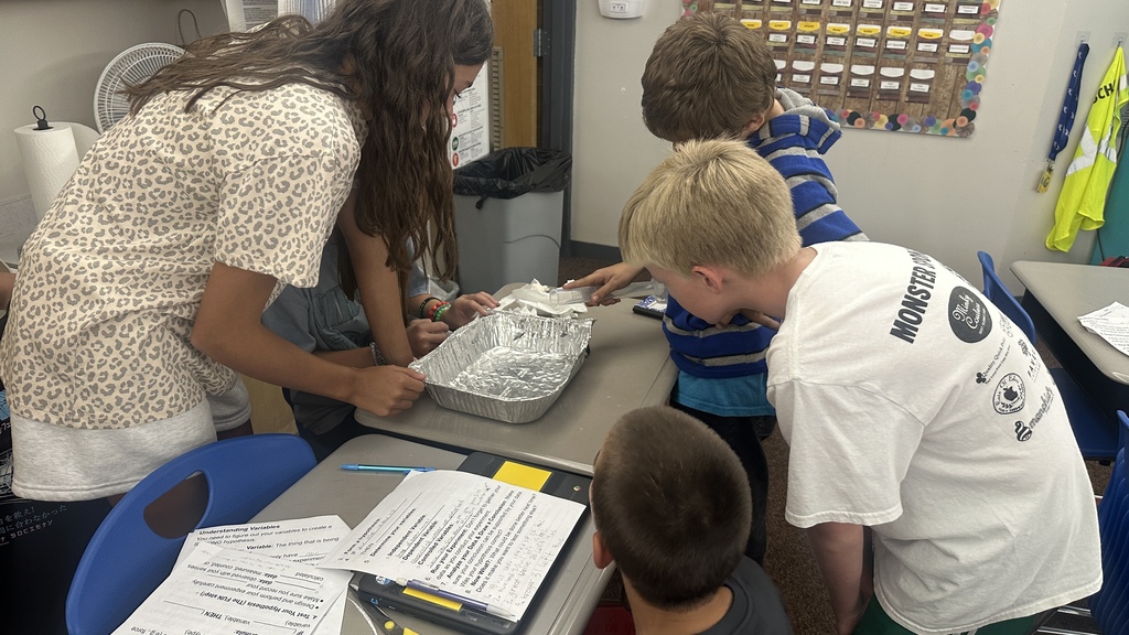 Students gathered around a desk using a clear graduated cylinder and an aluminum pan during a hands-on classroom lab. 