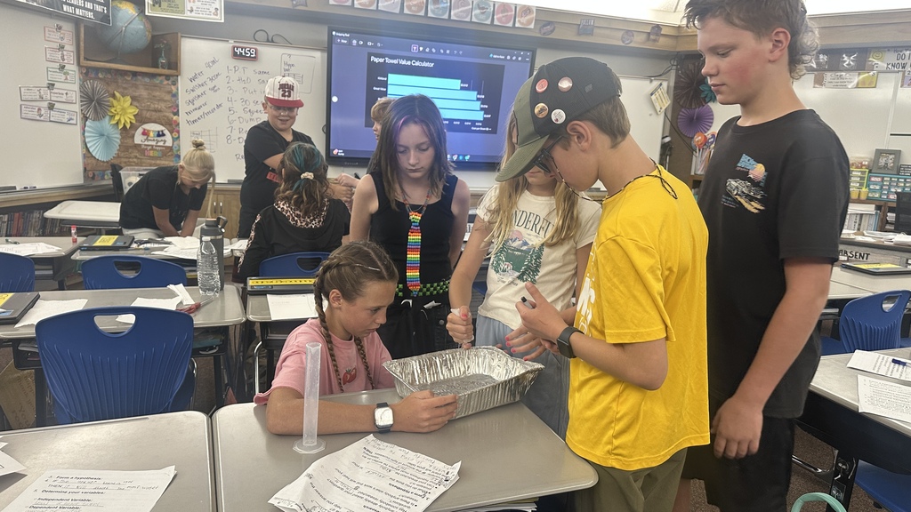 Students collaborating around a table with a foil pan, measuring cylinder, and lab worksheets, with a data chart visible on a screen in the background.  