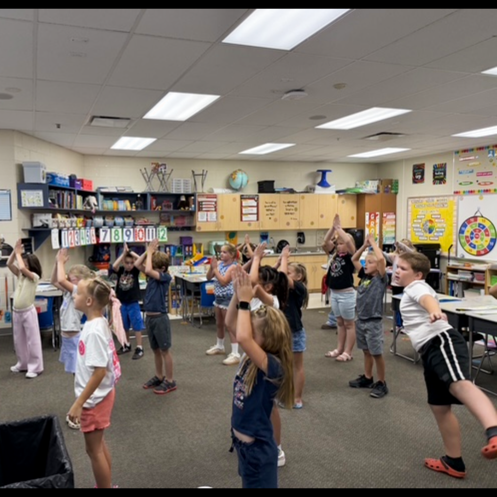 A class of students standing with their hands above their heads in their classroom.