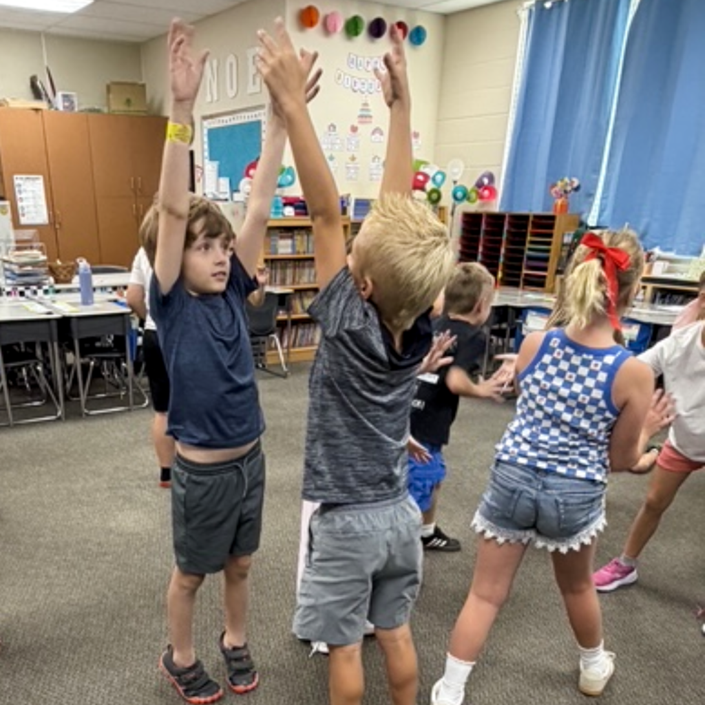 A class of students standing in their classroom stretching as high as they can.